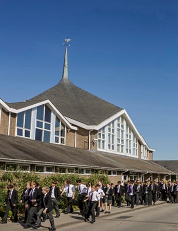 School building with a spire topped by a dove weathervane; many students in uniform walk past on a sunny day.